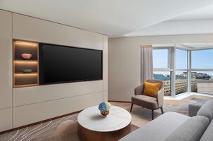 Una habitación de hotel moderna con una amplia televisión de pantalla plana montada en la pared, un sofá cómodo, un sillón pequeño con un cojín amarillo y una mesa de centro redonda con un jarrón.