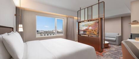 A hotel room with a large bed, a sofa, a TV, and a window with a city view.