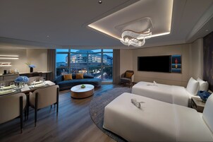 A modern hotel room with a dining area, a sofa, a TV, and a view of the cityscape.