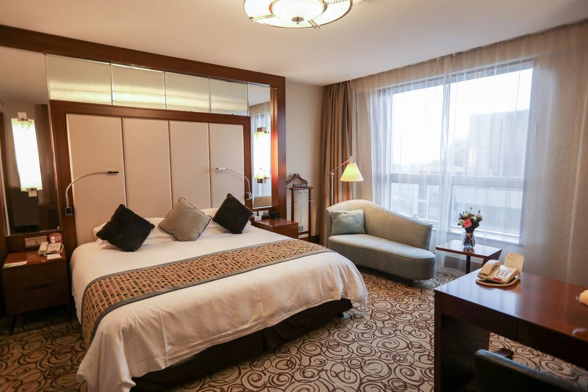 Una habitación de hotel con una cama grande, un escritorio, una silla, una lámpara y un amplio ventanal con cortinas.