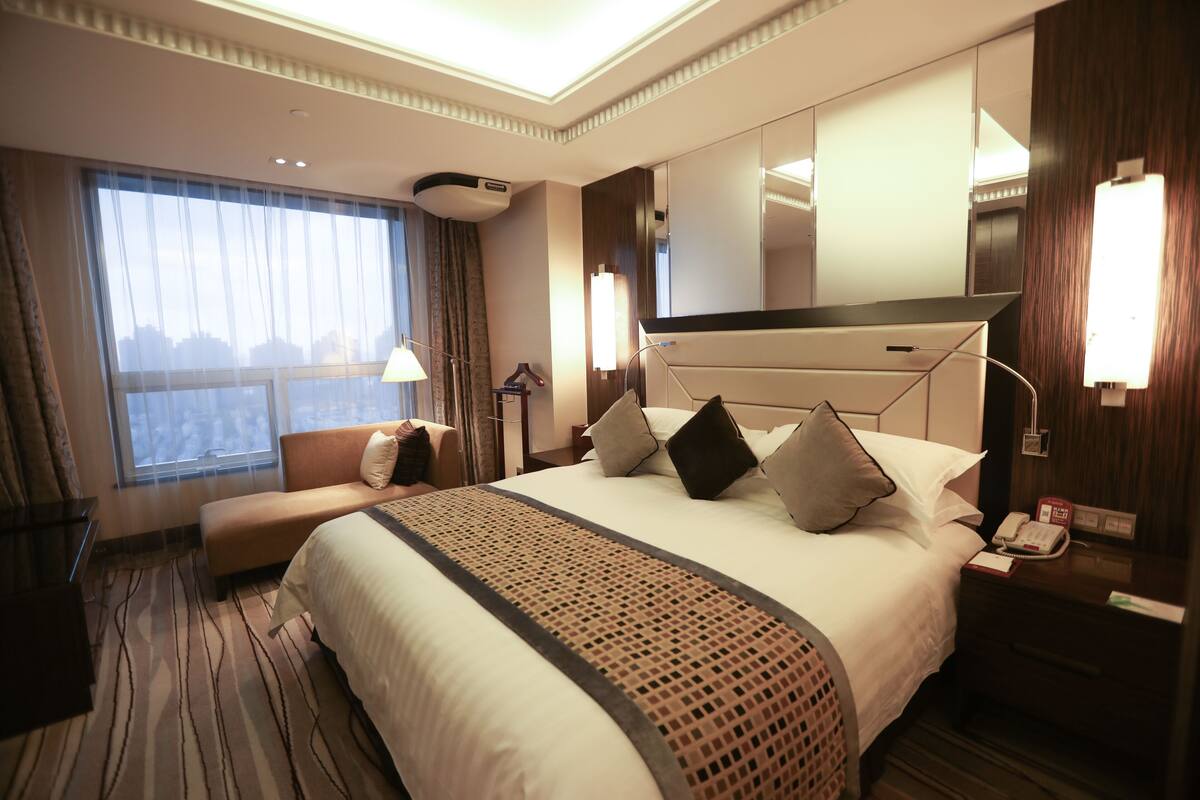 Habitación de hotel con una cama grande, una mesita de noche con teléfono, una lámpara y vistas a la ciudad a través de la ventana.