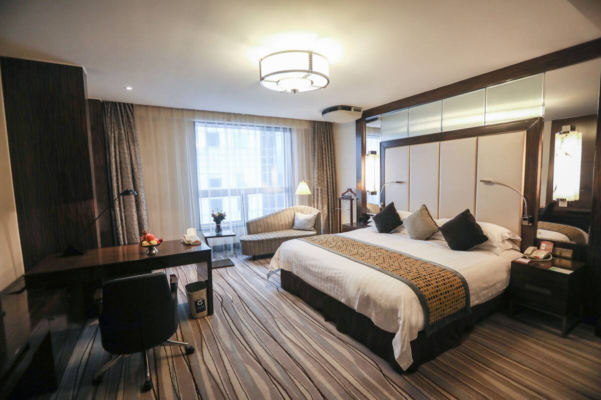 Habitación de hotel con una cama grande, un escritorio, una silla, una lámpara y un ventanal con cortinas.