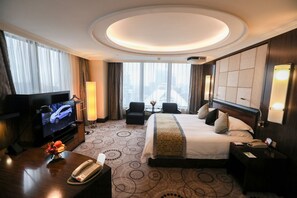 A hotel room with a large bed, a TV, a desk with a telephone, and a view of the city through large windows.