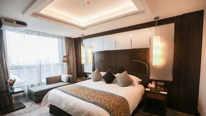 A hotel room with a large bed, a bedside table with a lamp, a nightstand with a phone, and a large window with curtains.