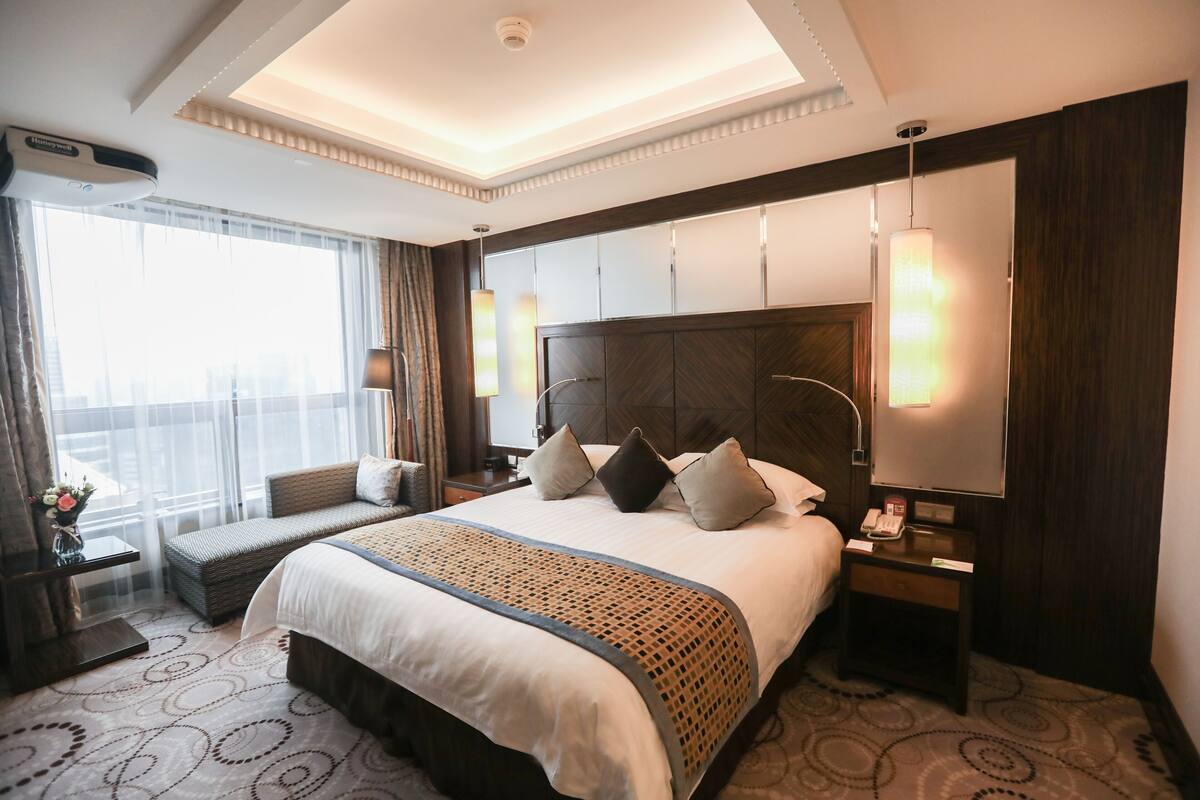 Habitación de hotel con una cama grande, una mesita de noche con lámpara, un aparador con teléfono y un ventanal con cortinas.