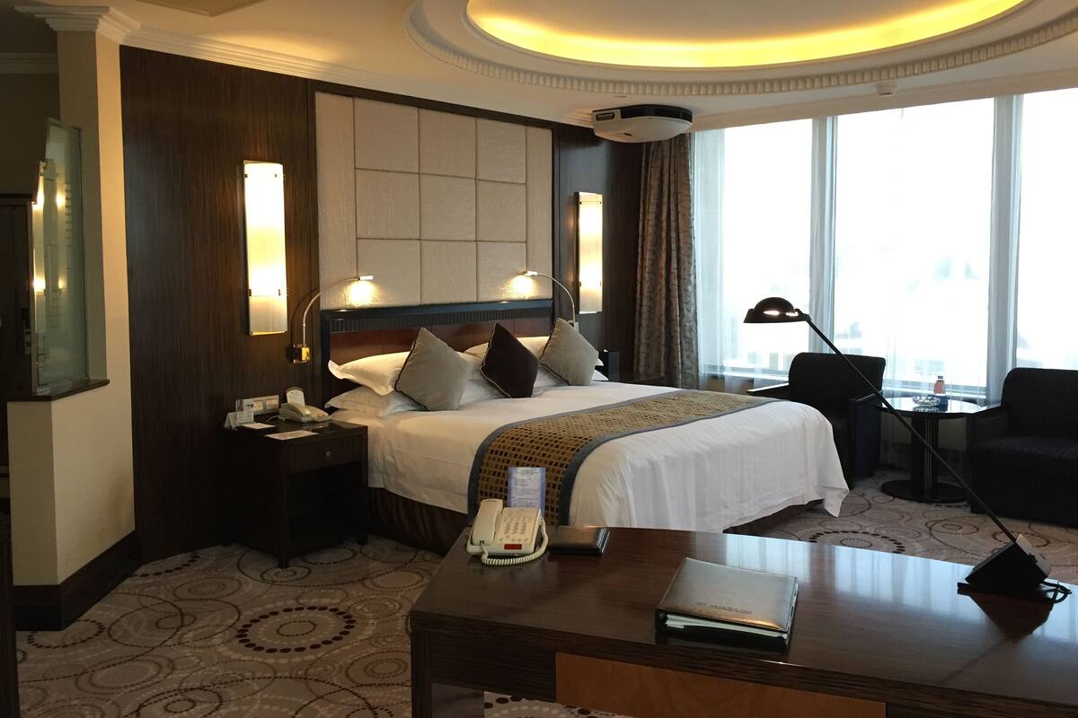Una habitación de hotel con una cama grande, lámparas de noche, un escritorio con teléfono y una carpeta, una silla y un amplio ventanal con cortinas.