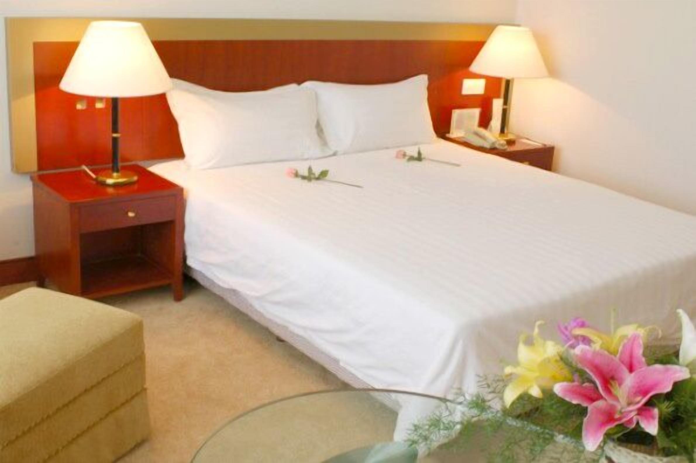 A hotel room with a large bed, two bedside tables with lamps, a small bedside table with a phone, a beige armchair, and a vase with flowers.