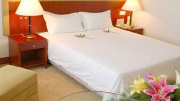 A hotel room with a large bed, two bedside tables with lamps, a small bedside table with a phone, a beige armchair, and a vase with flowers.