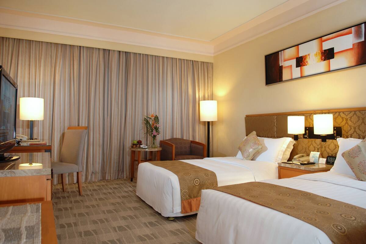 Quarto de hotel com duas camas, uma escrivaninha com luminária, uma cadeira, uma mesinha de centro com um vaso e um quadro na parede.