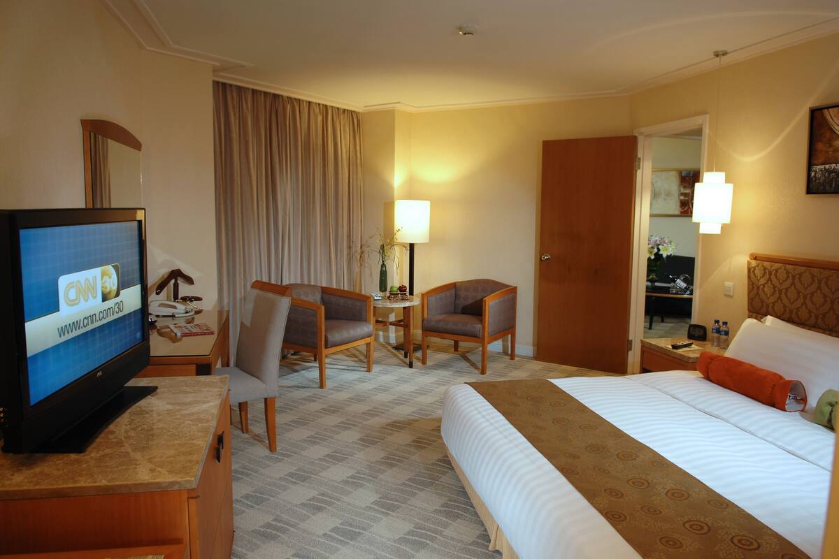 Quarto de hotel com cama, duas poltronas, televisão, abajur e escrivaninha com telefone.