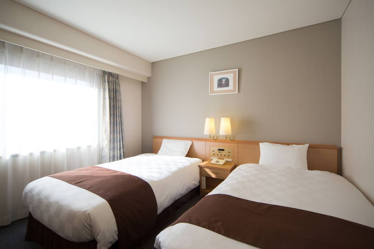 Een hotelkamer met twee bedden, een nachtkastje met lampen, een klok en een ingelijst schilderij aan de muur.