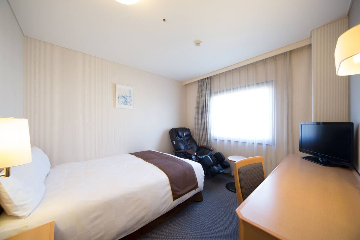 Een hotelkamer met een bed, een bureau met televisie, een stoel en een raam met gordijnen.