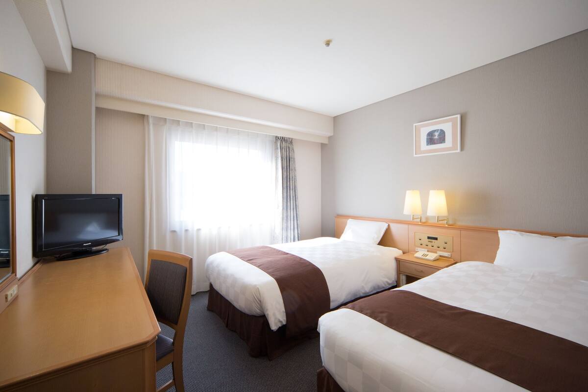 Een hotelkamer met twee bedden, een bureau met televisie, een nachtkastje met lampen en een raam met gordijnen.