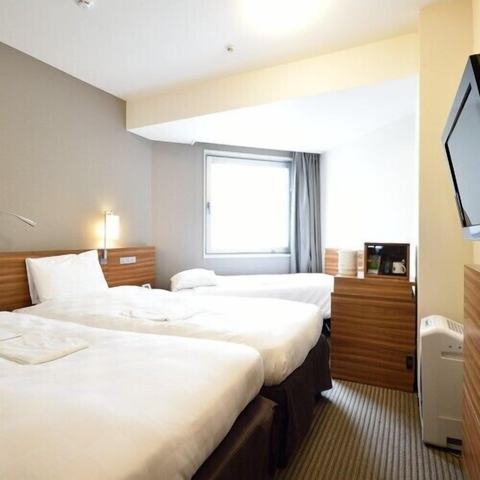 A hotel room with two beds, a TV, a window with curtains, and a bedside table with a lamp.