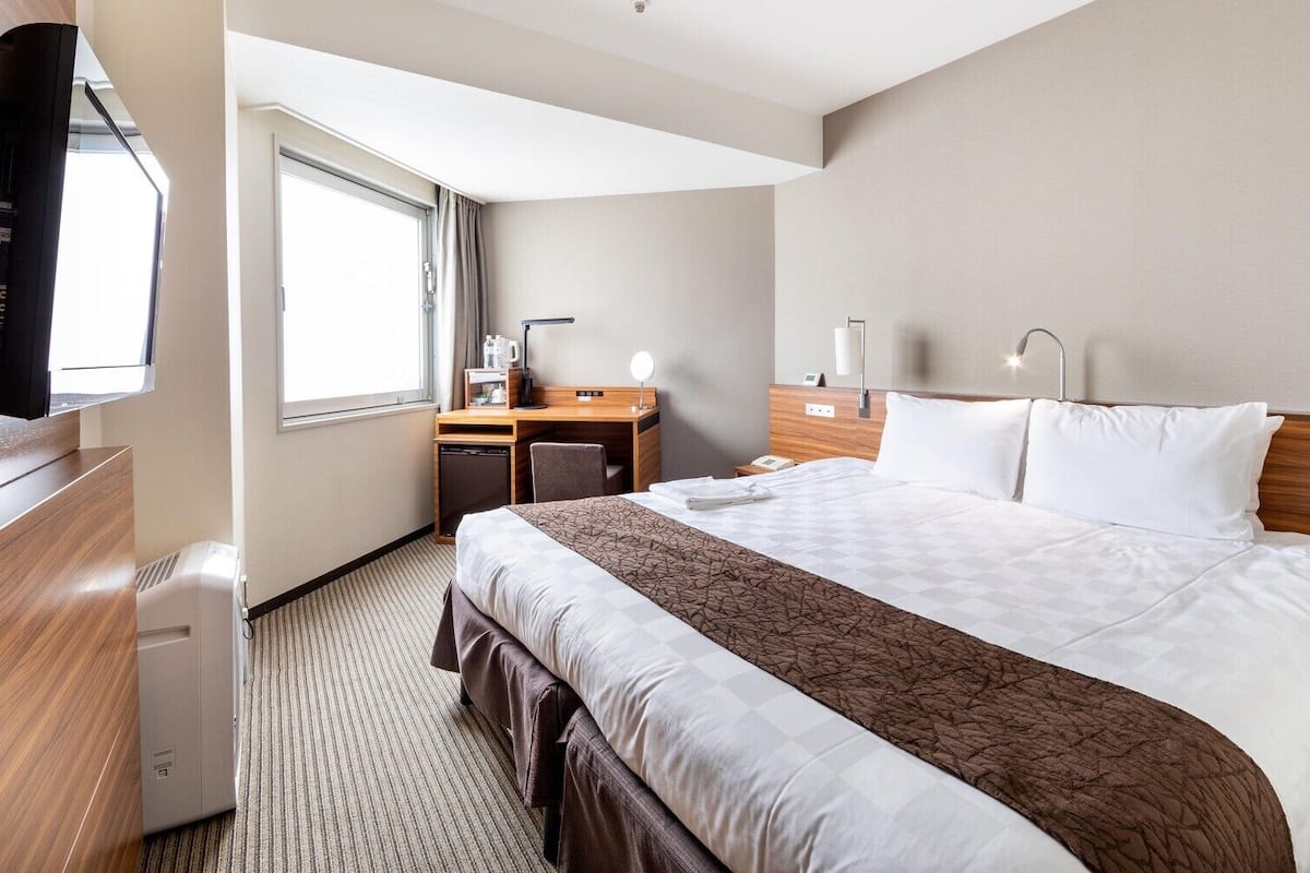 A hotel room with a bed, bedside tables, a desk with a chair, a window, and a television.