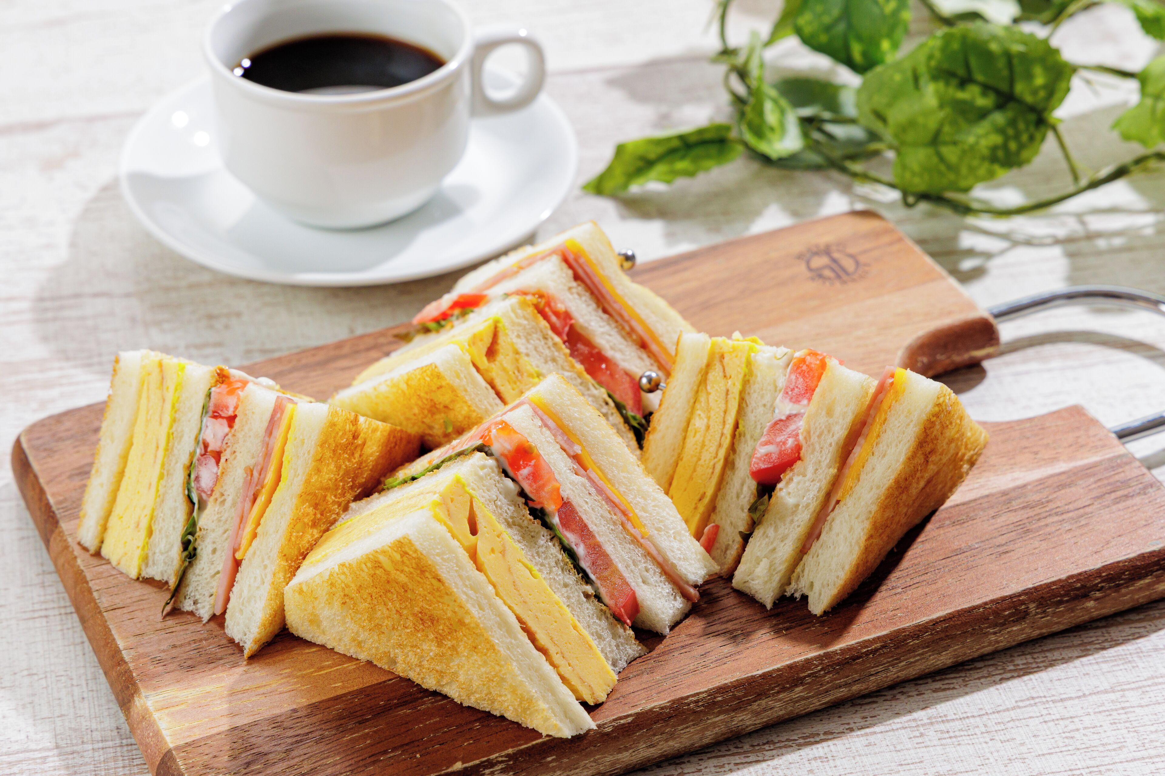 A sandwich cut into triangles on a wooden board, with a cup of coffee and green leaves in the background.