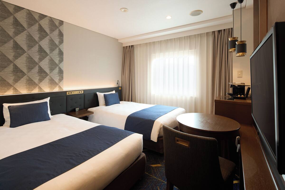 Hotelkamer met twee bedden, een bureau, een stoel, een televisie en een groot raam met gordijnen.