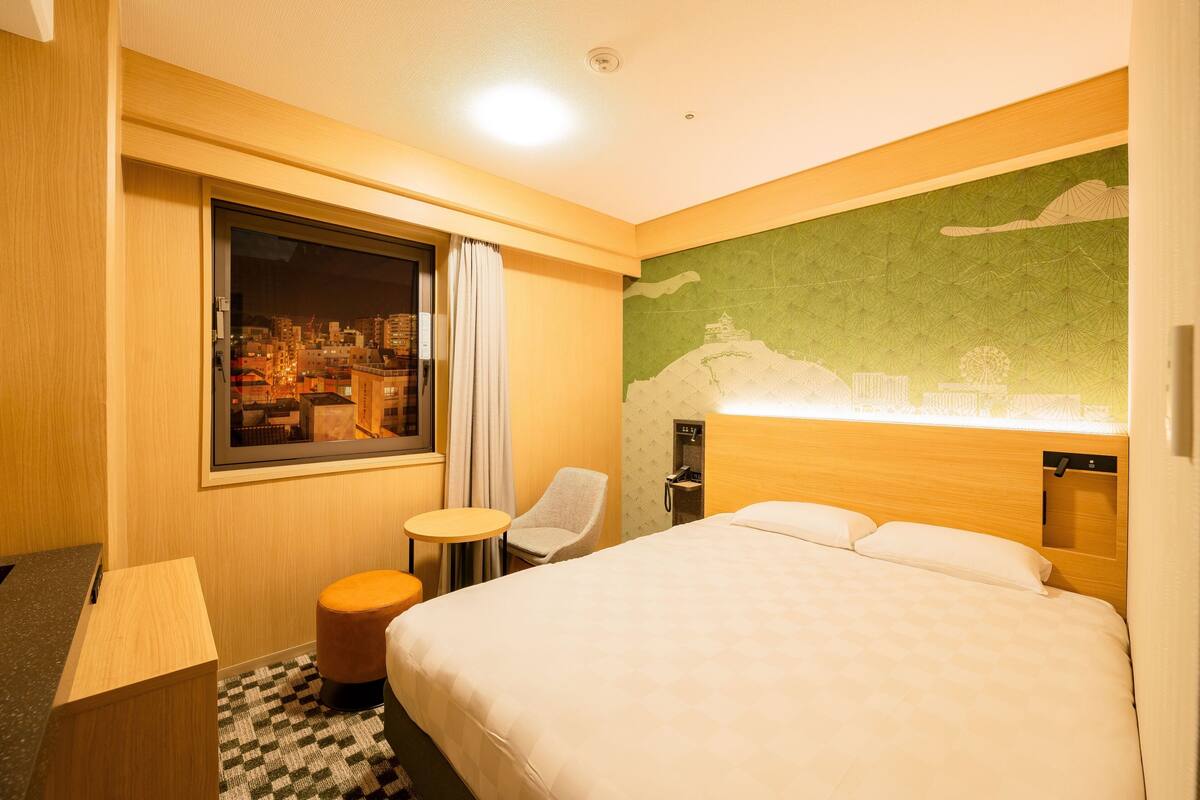 A hotel room with a large bed, a small table, a chair, a window with a city view, and a green wall with a cityscape mural.