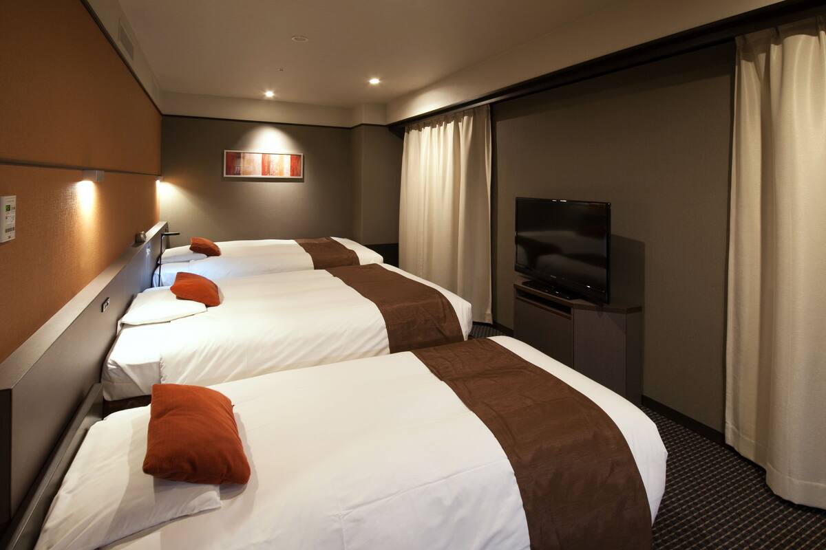 A hotel room with three beds, a flat-screen TV, and a large window with curtains.