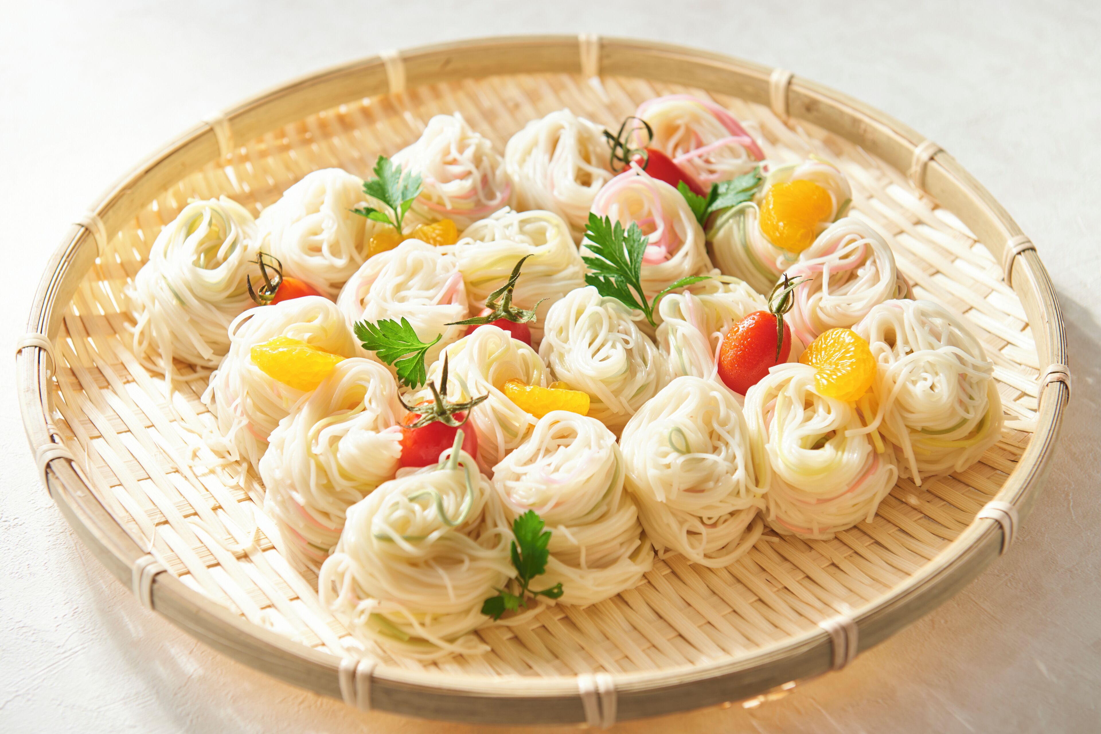A woven basket containing neatly arranged food items, likely a type of Asian noodle dish, garnished with herbs and small fruits.