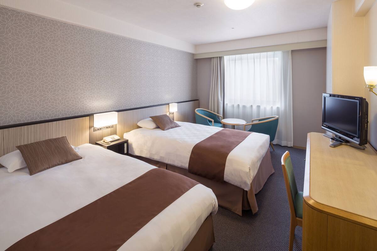 A hotel room with two beds, a desk with a television, a chair, and a window with curtains.