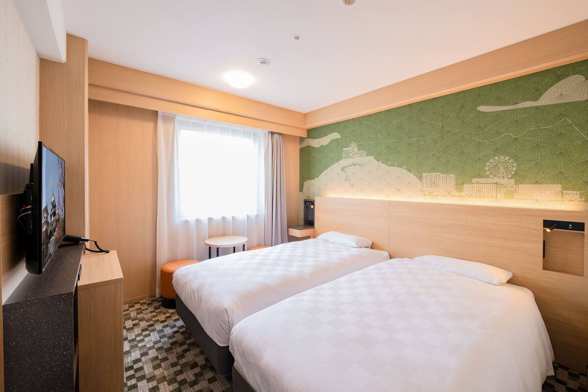 A hotel room with two beds, a desk with a TV, a small table, and a mural on the wall.