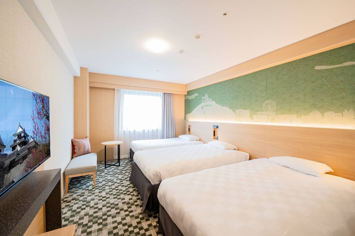 A hotel room with three beds, a TV, a small table, and a view of a cityscape.