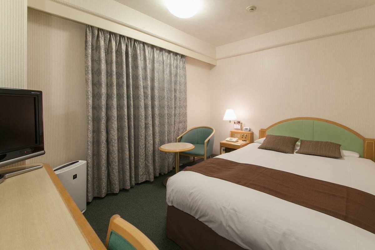 A hotel room with a bed, a desk with a television, a chair, a nightstand with a lamp, and a window with curtains.