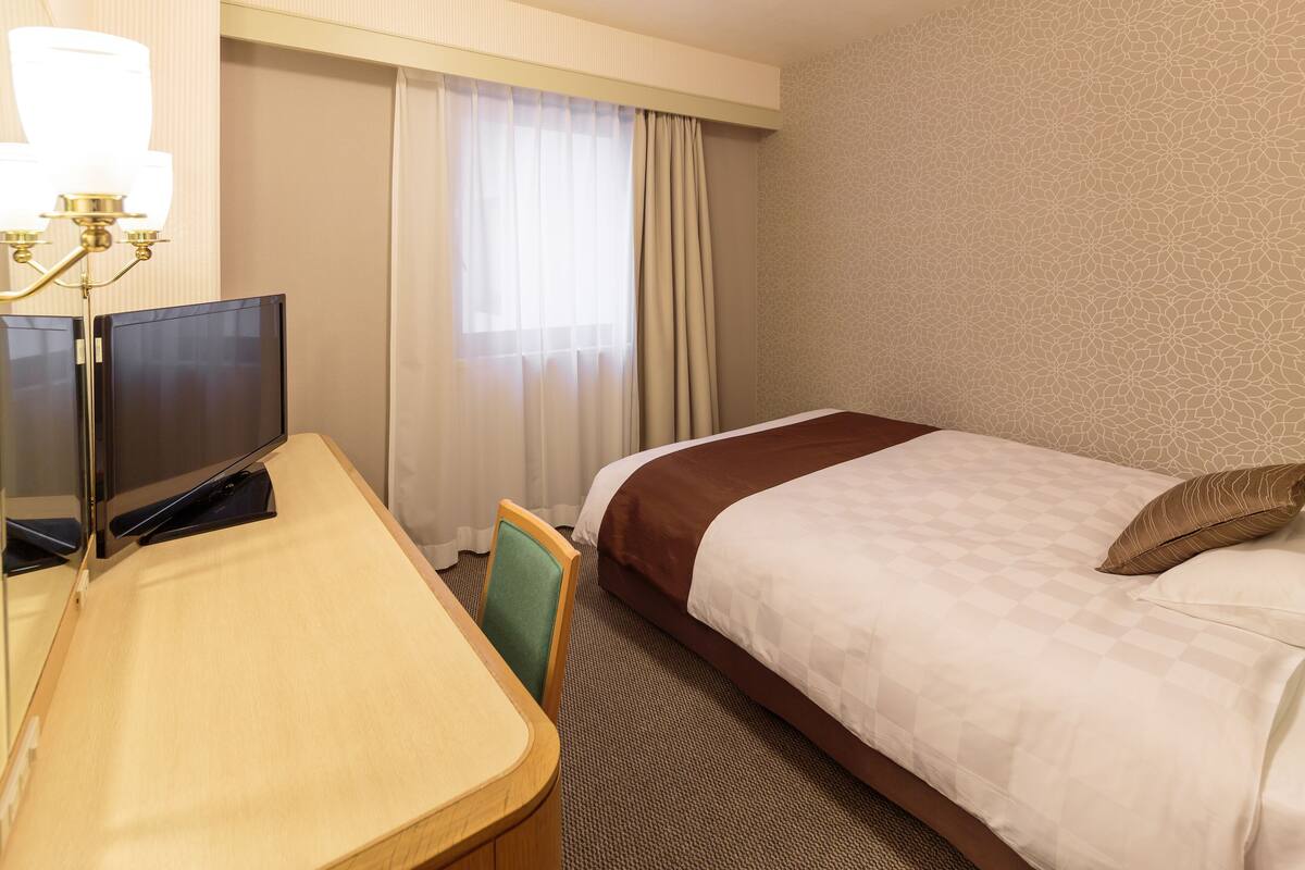 A hotel room with a bed, a desk with a television, a chair, and a window with curtains.
