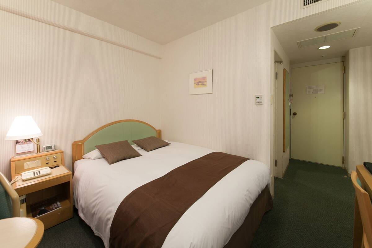A hotel room with a bed, bedside table, lamp, and a small desk with a telephone.