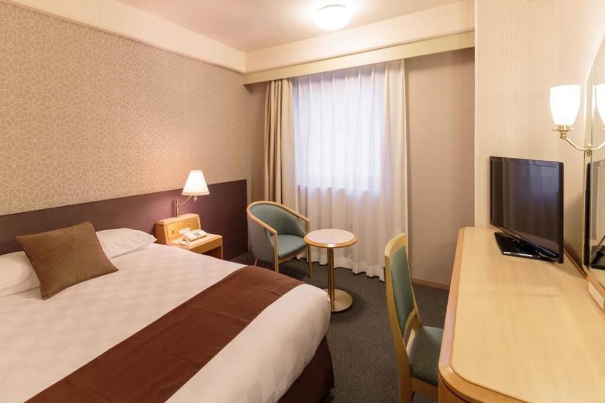 A hotel room with a bed, a desk, a chair, a TV, and a lamp.