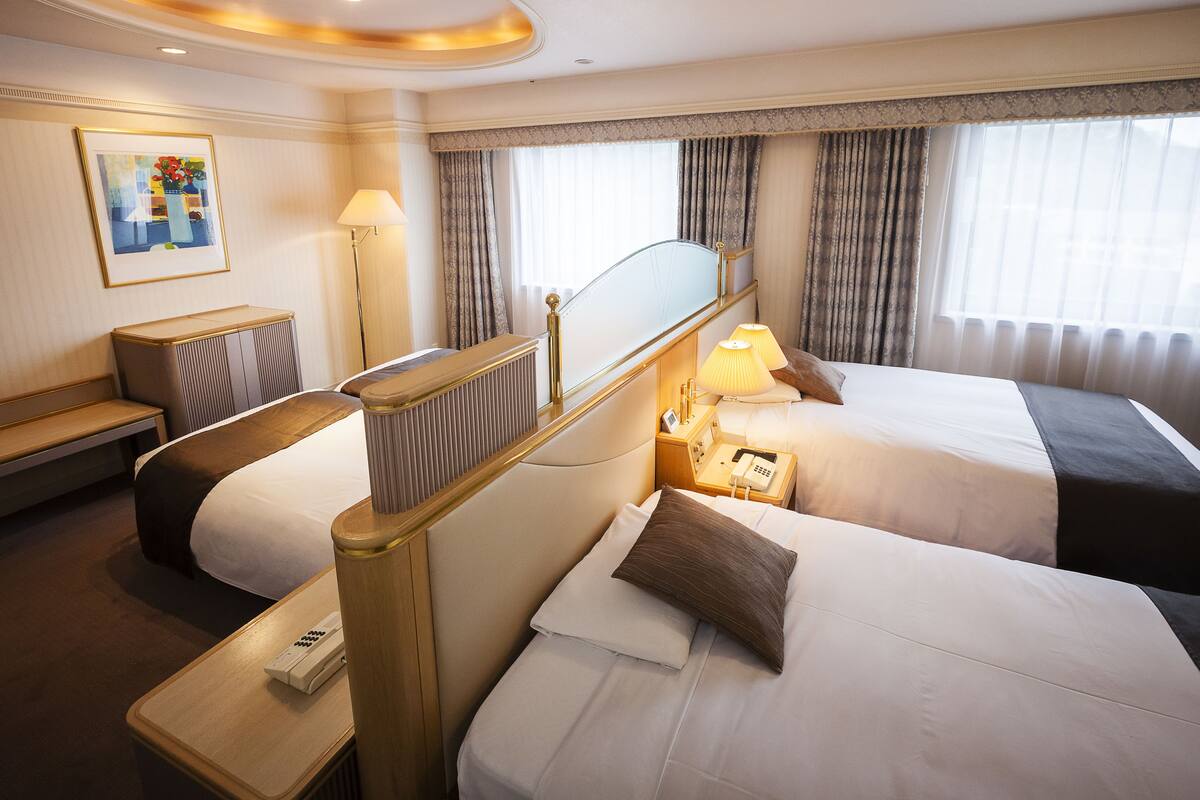 A hotel room with two beds, a bedside table with a lamp, a telephone, and a painting on the wall.