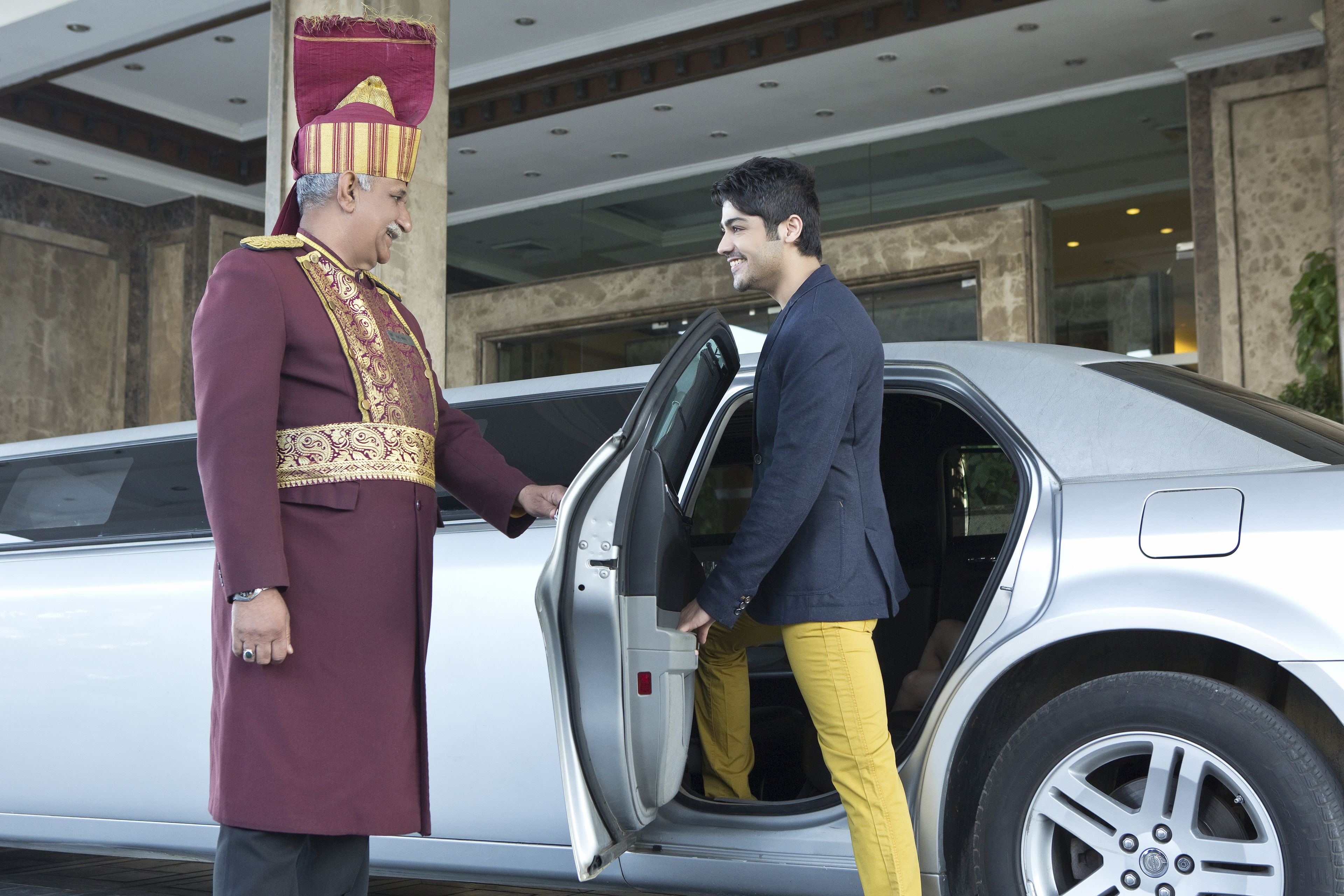 A man in a maroon uniform with a red and gold hat standing next to a silver car with its door open.