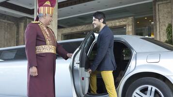 Un homme vêtu d’un uniforme bordeaux et coiffé d’un chapeau rouge et doré, se tient à côté d’une voiture argentée dont la portière est ouverte.