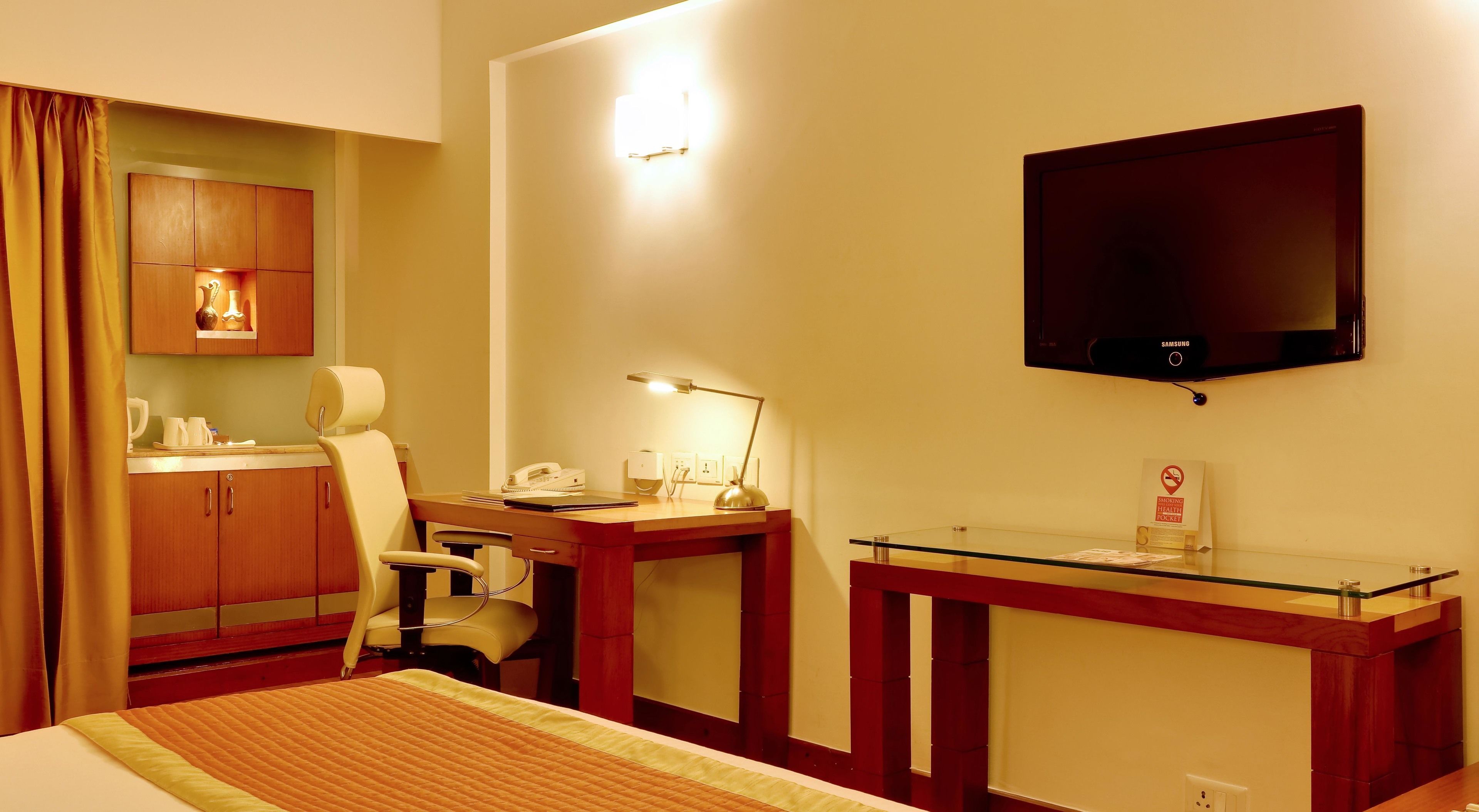 Habitación de hotel con cama, escritorio con lámpara, una silla, televisor y una mesita con superficie de vidrio.