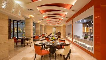 A modern restaurant with a central dining area featuring round tables and chairs, a kitchen visible through a glass partition, and a distinctive ceiling design with recessed lighting.