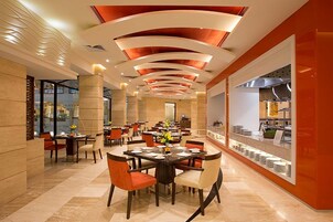 A modern restaurant with a central dining area featuring round tables and chairs, a kitchen visible through a glass partition, and a distinctive ceiling design with recessed lighting.