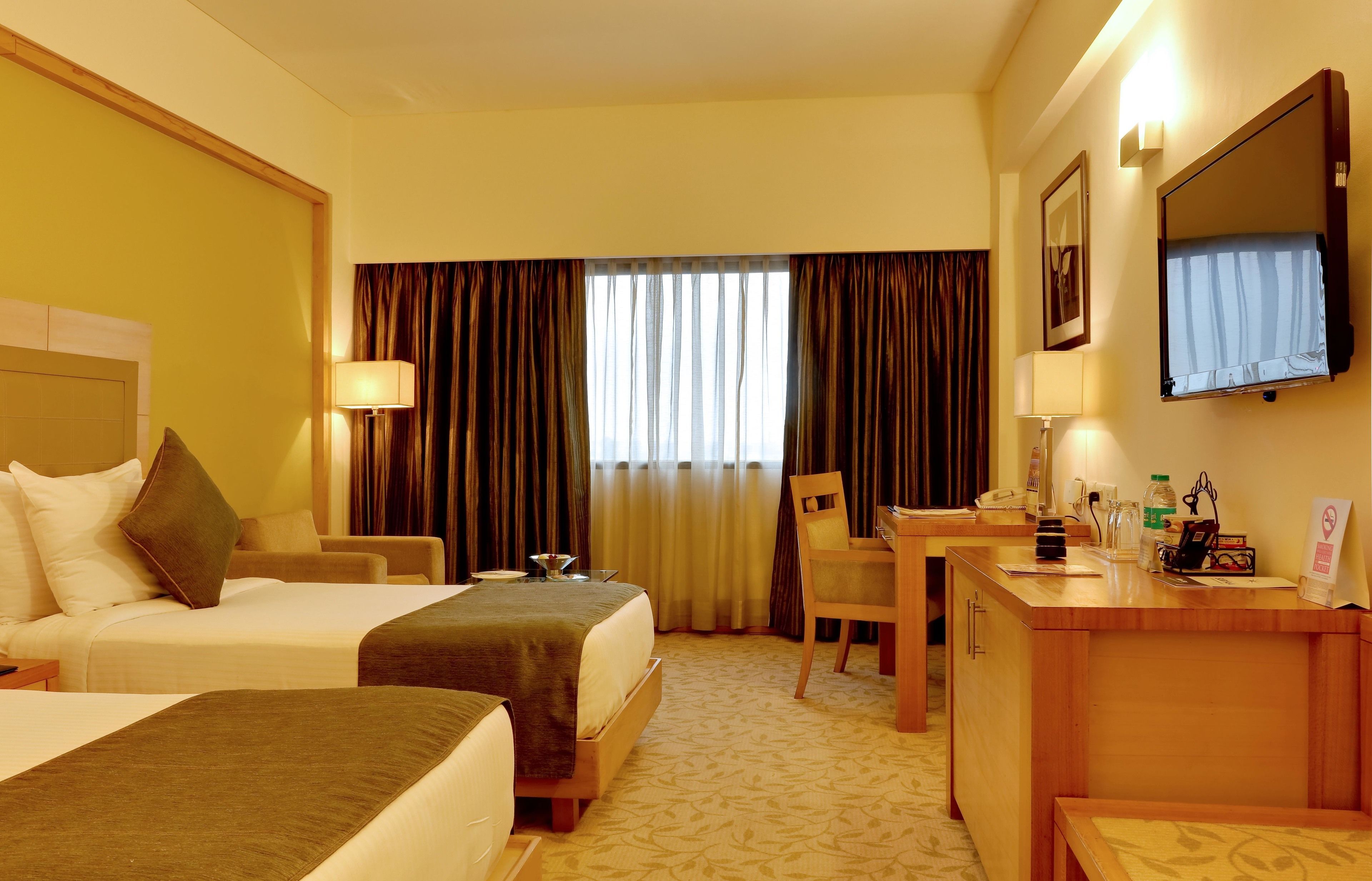 A hotel room with two beds, a desk, a chair, a TV, and a window with curtains.