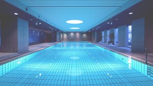 Une longue piscine rectangulaire intérieure, dotée d’un sol et de murs carrelés, entourée d’un couloir avec des luminaires encastrés et un plafond également carrelé.