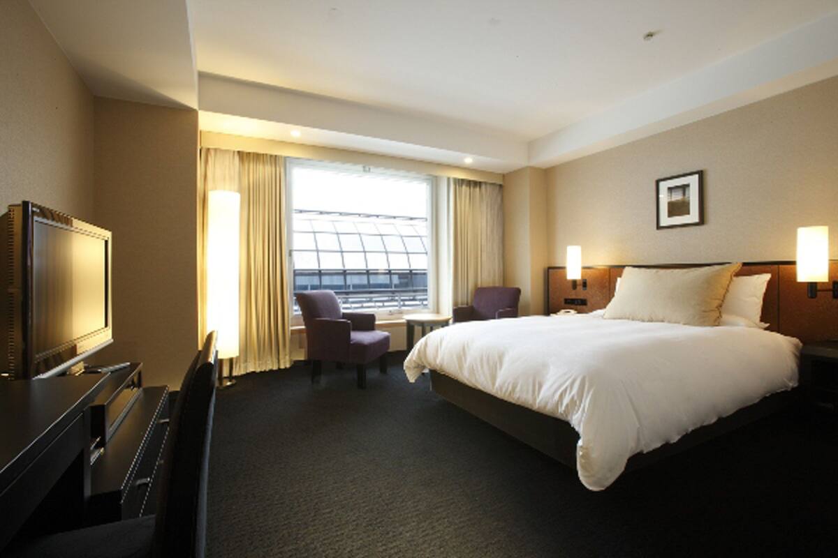 Una habitación de hotel con cama, escritorio con televisor, silla, mesita y ventana con cortinas.