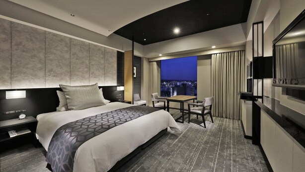 Une chambre d’hôtel moderne dotée d’un grand lit, d’un bureau avec des chaises, d’une télévision et offrant une vue sur la ville par la fenêtre.