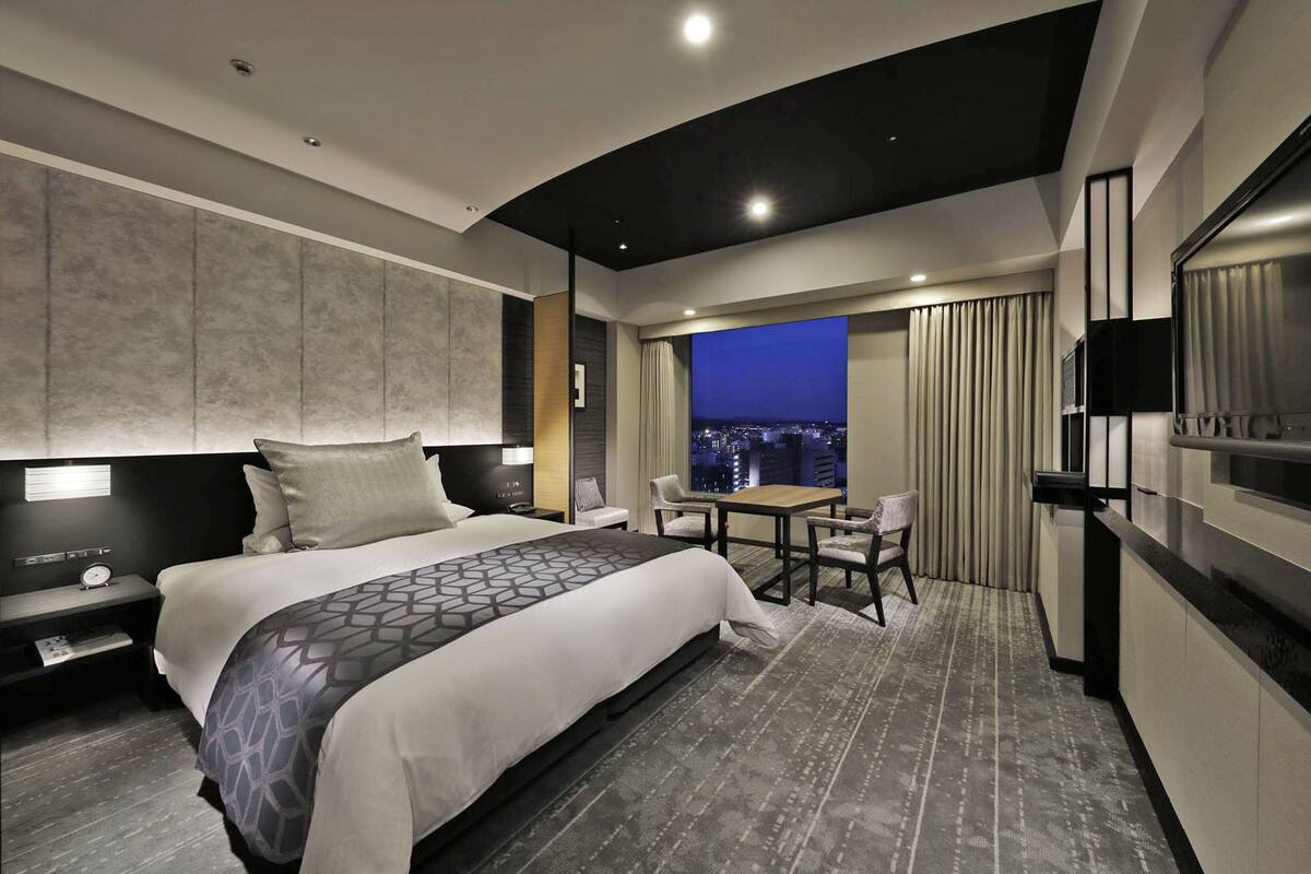 Een moderne hotelkamer met een groot bed, een bureau met stoelen, een televisie en uitzicht op de stad.
