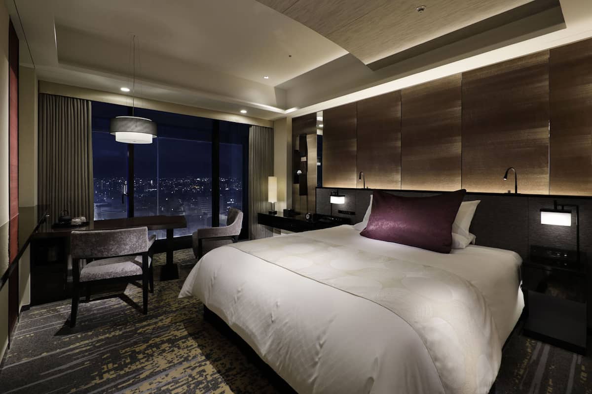 A modern hotel room with a large bed, a desk, and a chair. There is a city view through the window, and the room has a contemporary design with a wooden headboard and a large pendant light.