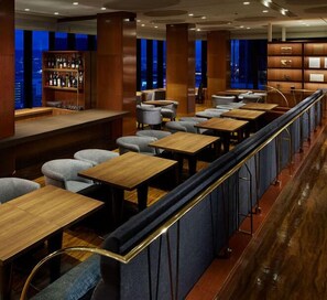 A restaurant with multiple tables and chairs, a bar area with shelves of bottles, and a view of the city at night.
