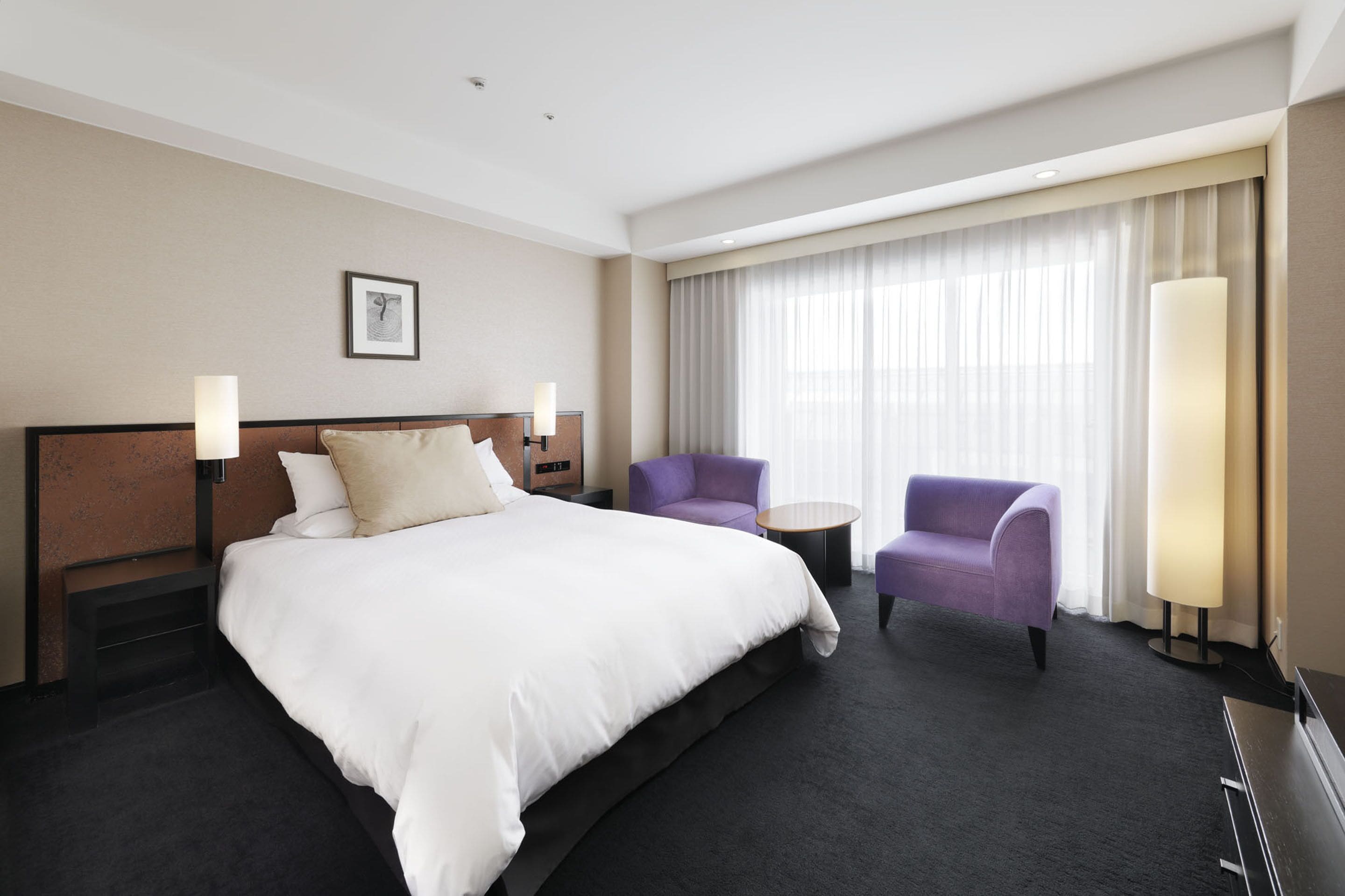 Habitación de hotel con una cama grande, dos sillones, una mesita y un ventanal con cortinas.