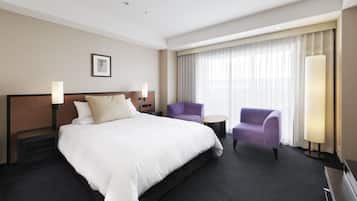 Habitación de hotel con una cama grande, dos sillones, una mesita y un ventanal con cortinas.