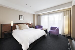 Habitación de hotel con una cama grande, dos sillones, una mesita y un ventanal con cortinas.