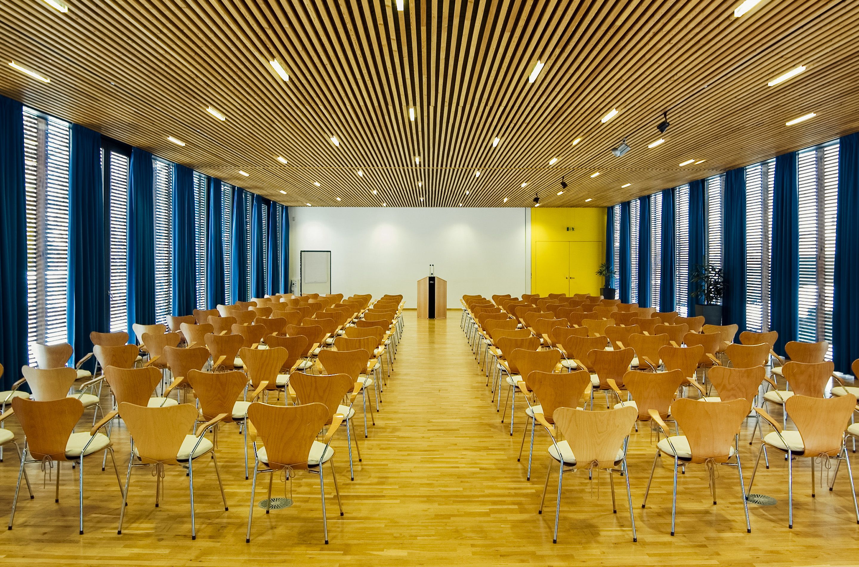 A conference room with rows of chairs, a podium, and large windows with blue curtains.