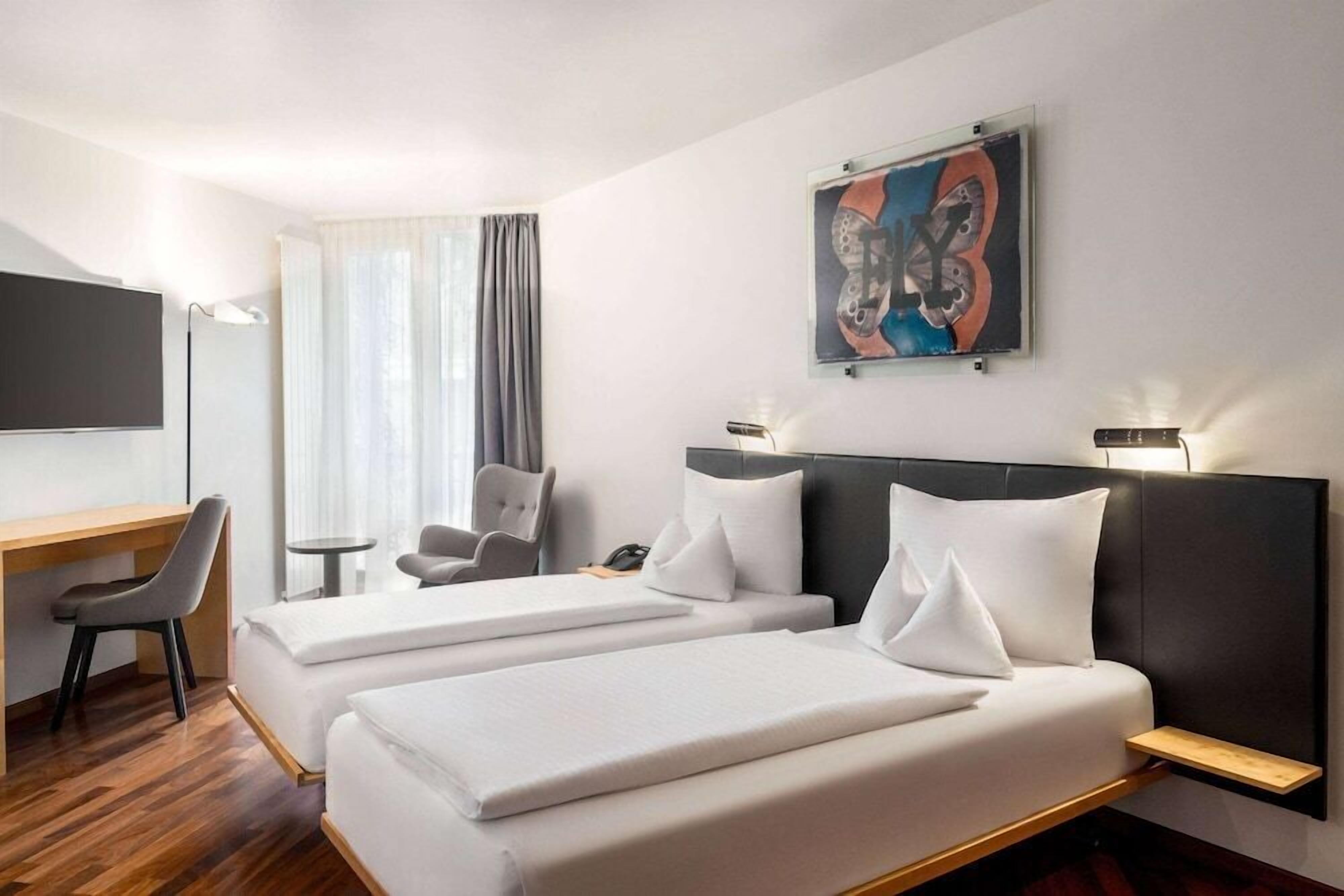 A hotel room with two beds, a desk, a chair, a TV, and a painting on the wall.