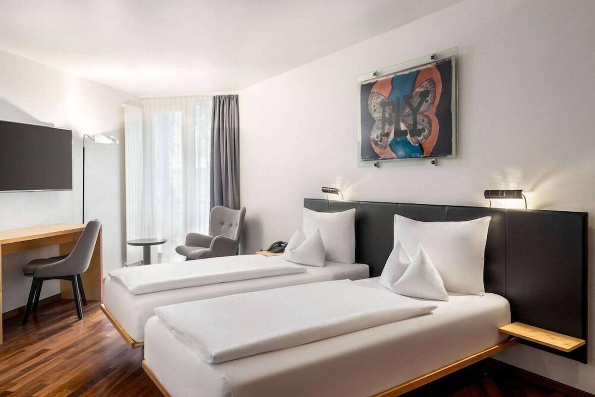 Quarto de hotel com duas camas, uma escrivaninha, uma cadeira, uma televisão e um quadro na parede.